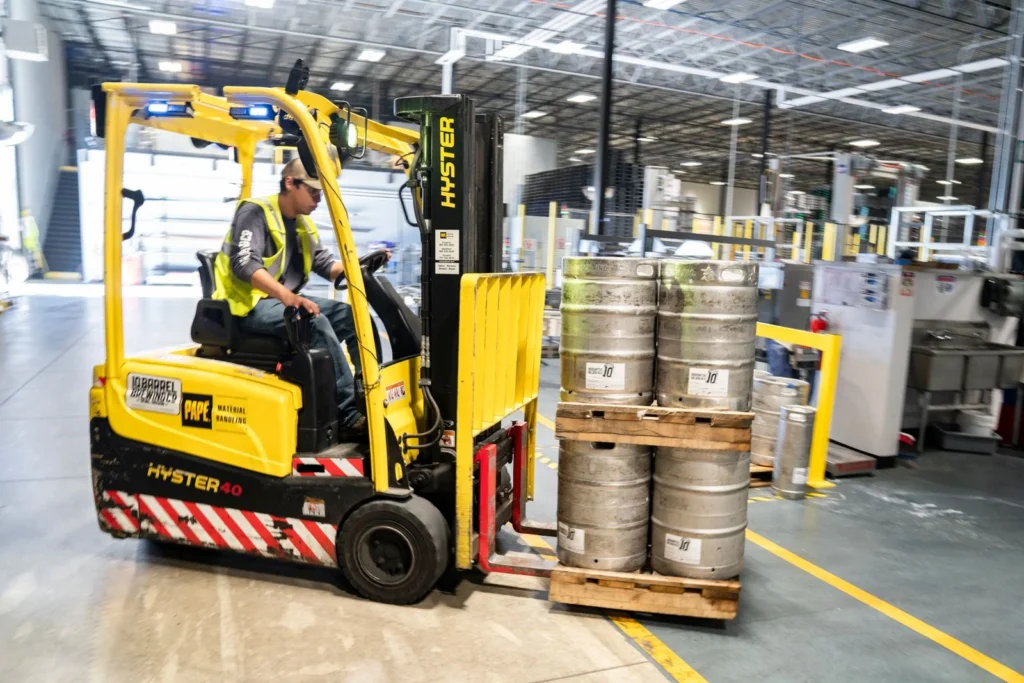 Forklift kuning mengangkat palet berisi tong logam di dalam gudang